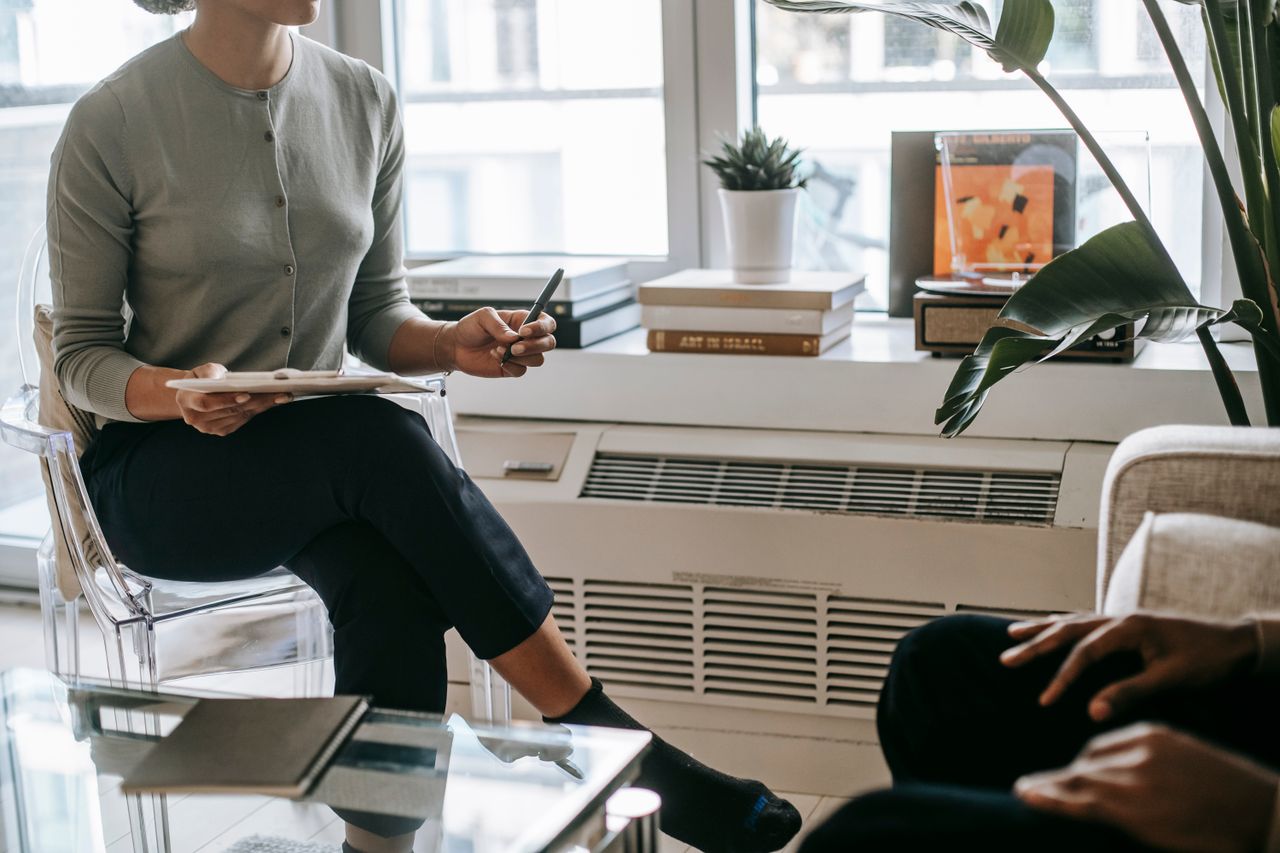 Unrecognizable female psychologist with crossed legs writing information on clipboard during appointment with anonymous ethnic patient in cozy light office
