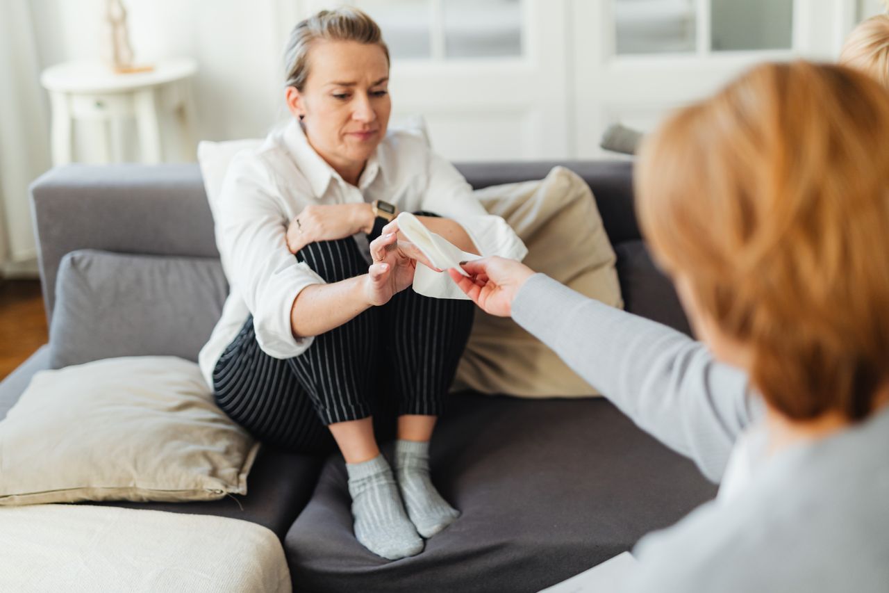 A woman consoles another with tissues, offering support in a comforting home setting.